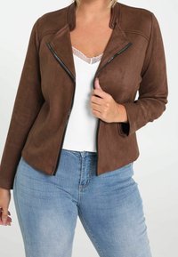 Nainen, joka on pukeutunut ruskeaan mokkatakkeeseen, valkoiseen pitsireunukseen yläosaan ja vaaleansinisiin farkkuihin, pitää takin vetoketjua rinnan lähellä.