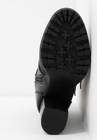 Semelle de botte en cuir noir avec un motif de bande de roulement profond comprenant des formes croisées et rectangulaires, inclinée contre l'angle du mur blanc et du sol.