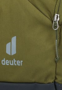 Zaino in tessuto verde oliva con zip argento, logo in evidenza e superficie testurizzata. Include un pannello inferiore grigio scuro.