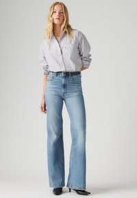 Jeans taille haute bleu clair avec un jambe droite, associés à une chemise blanche et rayée bleu marine à boutons. Ceinture noire et chaussures à bouts pointus.