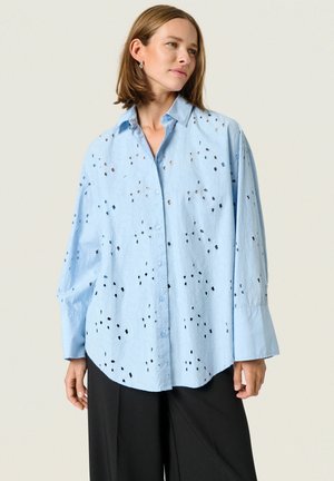  Lichtblauwe shirt met een losse pasvorm, voorzien van uitsnijdingen en een knoopsluiting aan de voorkant. Lange mouwen met brede manchetten en een afgeronde onderkant.