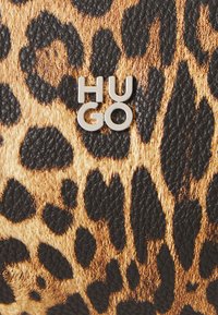 Leopardikuosis materiaali mustilla ja ruskeilla sävyillä, jossa on metallinen "HUGO" -logo näkyvästi esillä pinnalla.