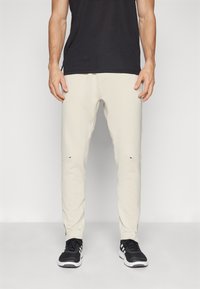 Beige sweatpants with a slim fit, featuring a textured surface, subtle side pockets, and contrasting dark accents; paired with black shoes.