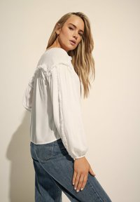 Hvid bluse med flæse detaljer langs ærmerne, med en løs pasform. Kombineret med højtaljede blå denim jeans. Glat tekstur.