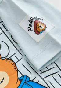 Hele sinine puuvillane T-särk, millel on multikakaru oranži näoga, sinist mantlit ja mütsi kandev ning õmmeldud sildiga "Paddington".