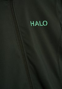 Close-up van donkergroene stof met zwarte rits en groen "HALO"-logo dat nabij de naad is gedrukt.