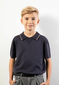 Blond chłopiec stojący na tle białej ściany, ubrany w ciemny sweter z krótkim rękawem z kołnierzykiem oraz szare spodnie we wzory z czarnym paskiem, ręce w kieszeniach.