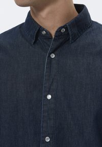 Camisa de mezclilla azul oscuro con cuello abotonado, acentos de botones plateados y una textura suave con costuras detalladas.