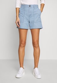 Femme portant un short rayé vertical bleu clair et blanc avec poches, une chemise blanche à manches longues et des baskets à semelles compensées blanches, debout les mains dans les poches.