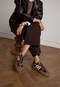 Braune Wildleder-Sneaker mit glitzernden Akzenten, runder Zehenpartie, dicke Sohle und kontrastierendem weißen Logo. Getragen mit dunklen, cropped Hosen.