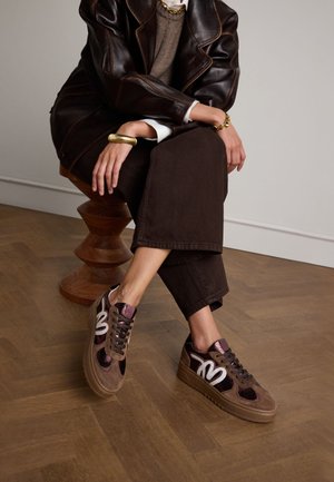 Bruine suède sneakers met glanzende accenten, ronde neus, dikke zool en contrasterend wit logo. Draag je met donkere, korte broeken.