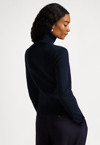 Ženska s temnimi kodrastimi lasmi nosi mornarsko modro majico z visokim ovratnikom in temne hlače, stoji bočno z rokami v žepih pred enobarvnim ozadjem.