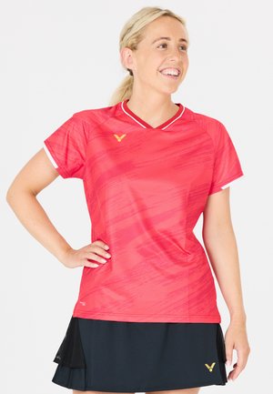 Smilende kvinde med blondt hår iført en rød kortærmet træningstrøje og sort sportsskjørt, stående med den ene hånd på hoften.