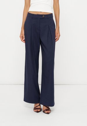 Femme portant un pantalon large bleu marine avec des plis à l'avant et un bouton marron, associé à des talons marron à brides et un top blanc.