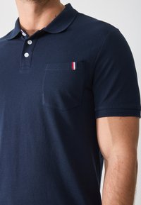Homme portant un polo bleu marine à manches courtes avec trois boutons blancs et une petite étiquette rouge, blanche et bleue sur la poche de poitrine.