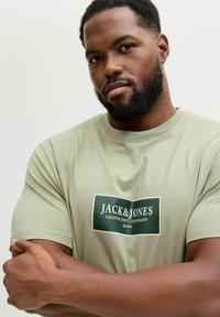 Camiseta de algodón verde claro con un estampado gráfico rectangular en verde oscuro con texto blanco, cuello redondo y mangas cortas.