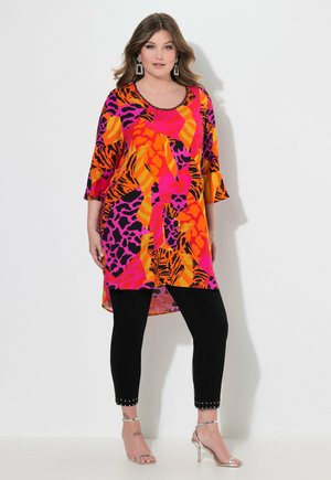 Helle, oversized Tunika in orange und pinkem Tierdruck, mit Dreiviertelärmeln und rundem Halsausschnitt. Kombiniert mit schwarzen Hosen.