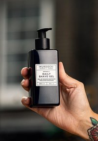 Svart, rektangulär flaska med pumpöppning, märkt "Daily Shave Gel" med vit text. Innehåller 250 ml gel. Hand greppande flaskan.