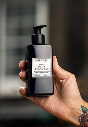 Bouteille rectangulaire noire avec un distributeur, étiquetée "Gel de rasage quotidien" en texte blanc. Contient 250 ml de gel. Main tenant la bouteille.