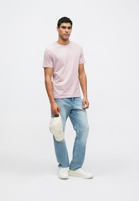 Svjetloružičasta pamučna majica s okruglim izrezom, uparena sa svijetloplavim trapericama. Model drži bijelu baseball kapu i nosi bijele tenisice.