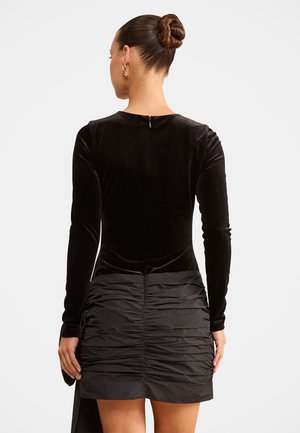 Femme avec les cheveux en chignon portant un haut en velours noir à manches longues et une mini-jupe noire froncée, debout de dos devant un fond uni.