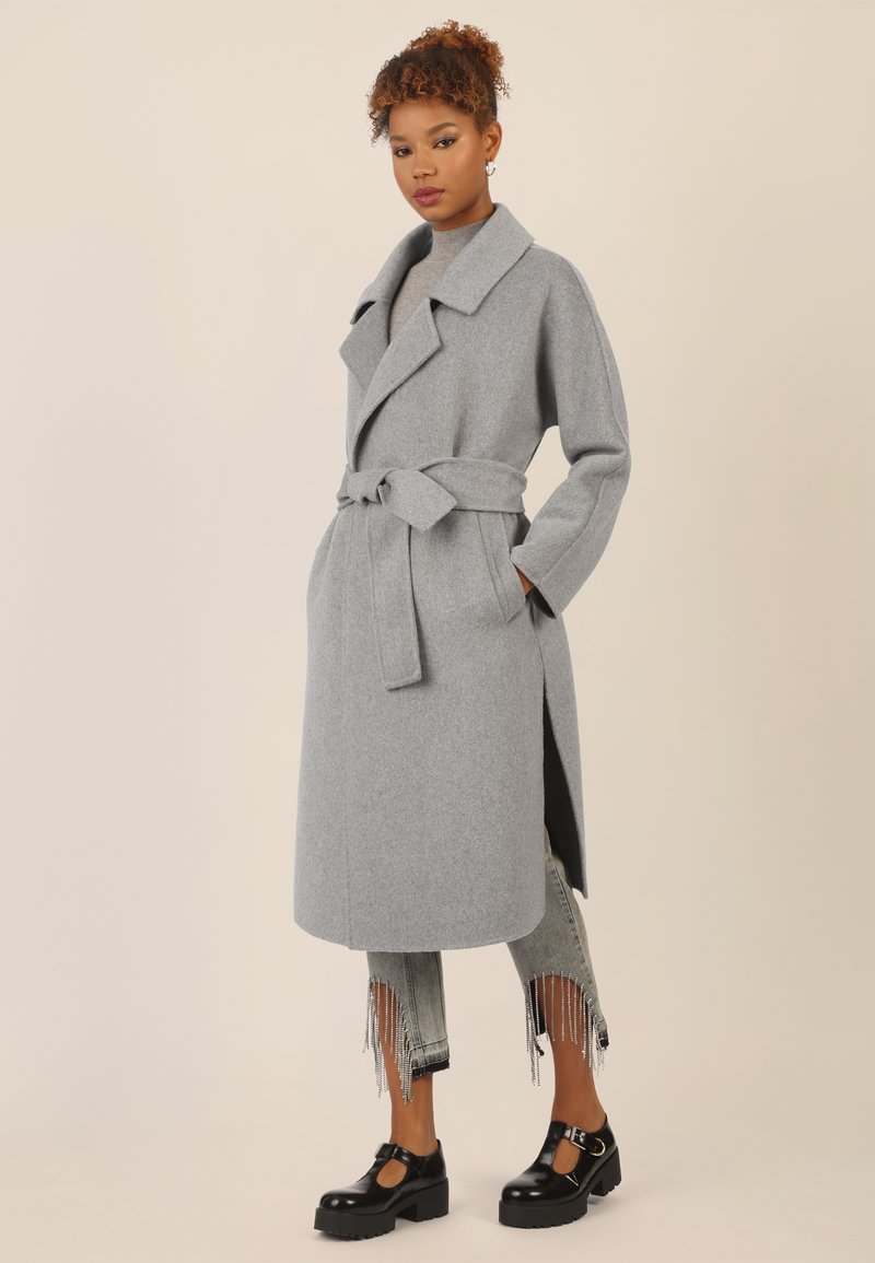 Cappotto di lana grigio con colletto a punta, vita cintata e fessure laterali, abbinato a jeans strappati e scarpe con plateau nere.