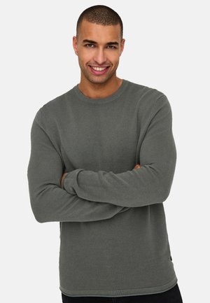 Uomo sorridente con capelli corti che indossa una camicia a maniche lunghe grigia e strutturata, in piedi con le braccia incrociate contro uno sfondo bianco semplice.