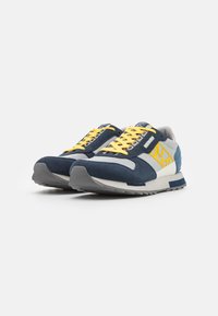 Sneakers blu e grigi con tomaia in suede e mesh, lacci gialli e suola in gomma con un motivo testurizzato. Dettaglio del logo ben visibile sul lato.