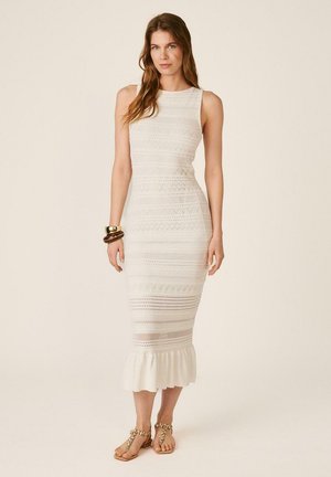 Mujer con vestido midi blanco sin mangas con patrones de encaje y dobladillo con volantes, complementado con pulseras doradas y sandalias decoradas.