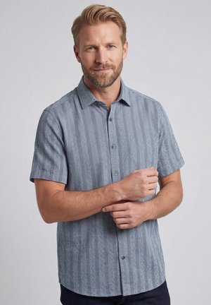 Blond man met baard die een blauw gestreept overhemd met korte mouwen draagt, poseert met één hand terwijl hij de andere arm aanpast tegen een effen achtergrond.