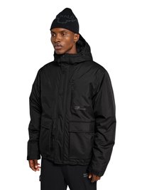 Mann trägt eine schwarze Winterjacke mit mehreren Taschen, einer Kapuze und einer schwarzen Strickmütze, steht vor einem weißen Hintergrund.