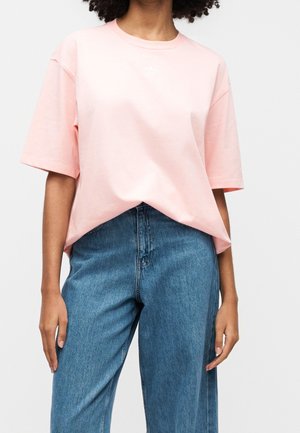 Personne portant un t-shirt ample à manches courtes rose clair et un jean bleu taille haute, debout, les bras détendus le long du corps.
