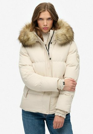 Beige pufferjack met een bont afgewerkte capuchon, gequilte ontwerp, klepzakken en geribde manchetten. Het materiaal lijkt zacht en warm.