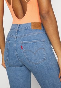 Kvinna som bär högmidjade blå Levi's-jeans och en persikofärgad ärmlös topp med låg rygg, som visar bakfickorna och varumärkets läderpatch.