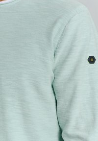 Licht mintgroene gebreide trui met een ronde hals en een zwart hexagonaal logopatch op de linker mouw. Zachte textuur, casual ontwerp.