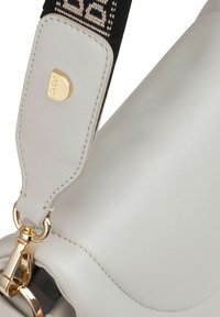 Close-up van een lichtgrijze leren tas met een gouden sluiting en een zwarte strap, voorzien van beige tekstdetails.
