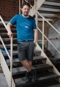 Camiseta azul con un gráfico geométrico de montañas, pantalones cortos cargo grises y botas negras con cordones. De pie en las escaleras con una mano en la barandilla.