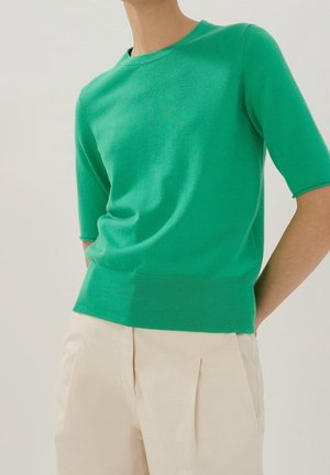 Persoon die een aansluitend groen shirt met korte mouwen en beige, hooggetailleerde geplooide broek draagt tegen een effen achtergrond.