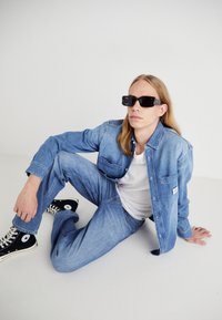 Giacca in denim e pantaloni in blu chiaro con una maglietta bianca e sneakers nere. La giacca ha una tasca e chiusura con bottone, indossata in modo informale.