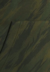 Grön camouflagemönstrad tyg med varierande nyanser och texturer, med en synlig fick söm, lämplig för avslappnad klädsel eller utomhusaktiviteter.