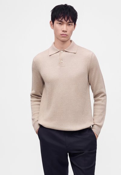 Jeune homme aux cheveux noirs portant un pull polo beige à manches longues et un pantalon sombre, debout, les mains dans les poches, devant un fond uni.