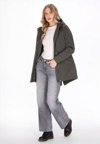 Femeie purtând o jachetă cu glugă de culoare verde măsliniu peste o cămașă albă, blugi gri cu picioare largi și cizme negre, stând cu o mână în buzunar.