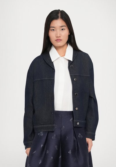 Woman with long dark hair wears a white collared shirt, dark denim jacket with buttons, and navy skirt with subtle floral patterns.