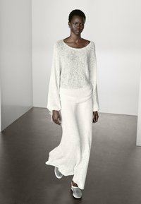 Maglione bianco a maglia con maniche ampie, abbinato a una lunga gonna coordinata. La texture sembra morbida; la modella indossa scarpe slip-on di colore chiaro.