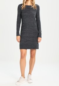 Abito lavorato a maglia grigio scuro con maniche lunghe, scollatura rotonda e forma aderente, abbinato a sneaker bianche per un look casual.