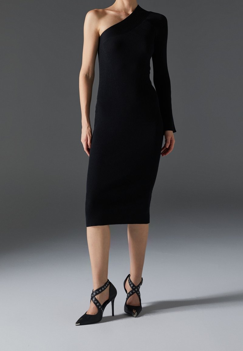 Femme portant une robe midi noire asymétrique à manches longues, associée à des chaussures à talons noirs avec des brides croisées.