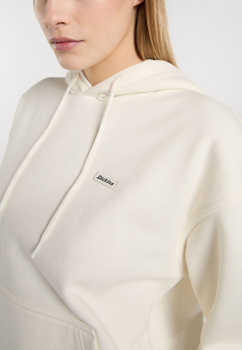 Sudadera color crema hecha de una tela suave, con un bolsillo frontal y una pequeña etiqueta de marca en el pecho. Cuerdas en la capucha.