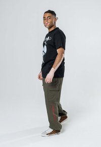 T-shirt grafica nera con design di teschio, pantaloni cargo verde oliva e scarpe con stampa leopardata. Accenti rossi ricamati notevoli sui pantaloni.