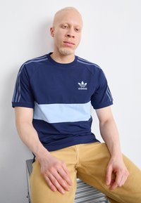 Polo majica s plavim i svijetloplavim prugama s Adidas logotipom; kratki rukavi, mekani pamučni materijal; nosi se s bež hlačama.