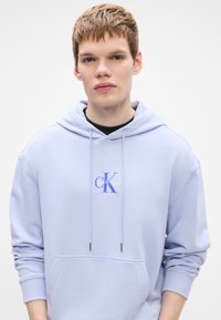 Svetlofialová mikina vyrobená z jemného materiálu s predným vreckom, s potlačeným logom v modrej a bielej farbe. Šnúrky s kovovými koncami.